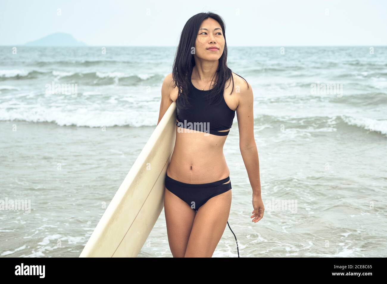Jeune femme asiatique surfeuse marchant sur une plage de sable et portant planche de surf sur mer bleu calme en regardant loin Banque D'Images