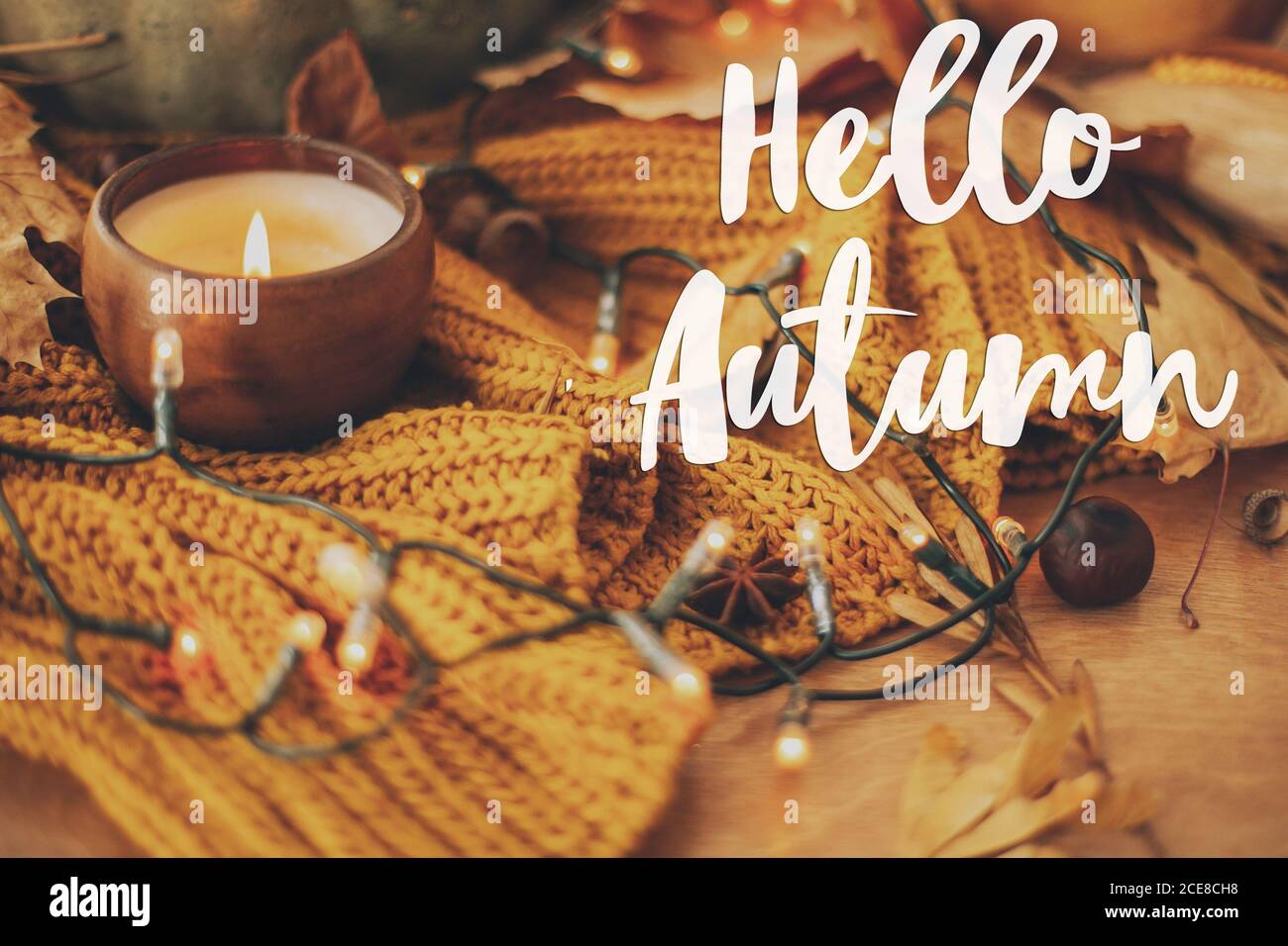 Carte de vœux Hello Autumn. Texte Hello automnal écrit à la main sur fond de citrouille, feuilles d'automne, bougie, lumières chaudes sur chandail tricoté jaune Banque D'Images