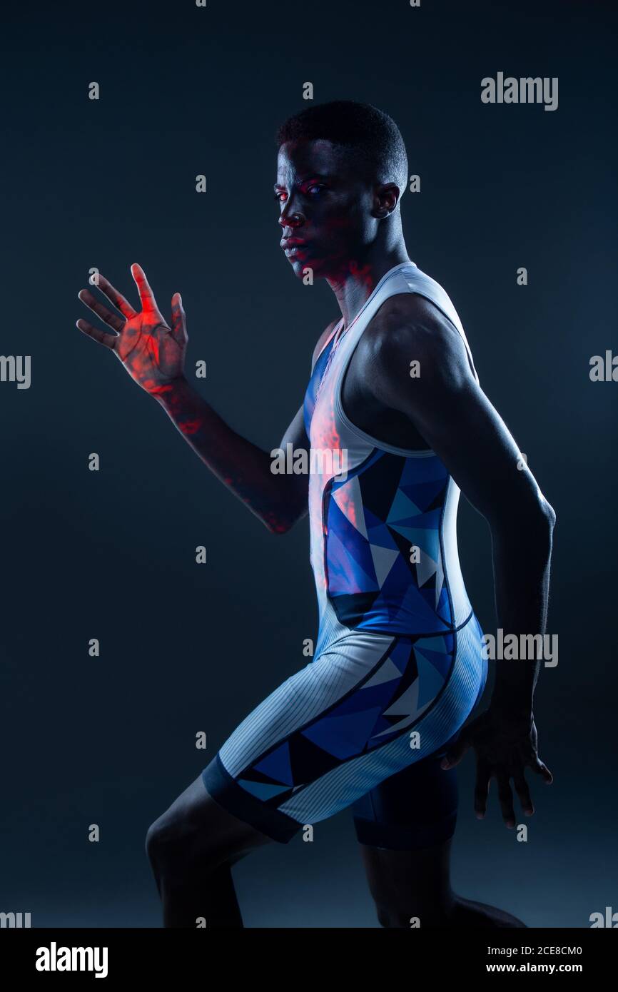 Vue latérale d'un sportif afro-américain sérieux et sportif en couleurs active wear pour des mouvements rapides en studio sombre avec éclairage au néon rouge Banque D'Images