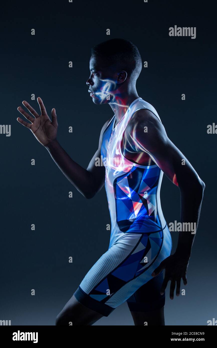 Vue latérale d'un sportif afro-américain sérieux et sportif en couleurs active wear pour des mouvements rapides en studio sombre avec éclairage au néon rouge Banque D'Images