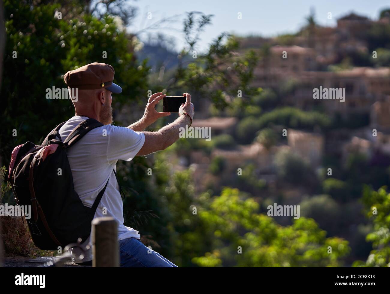 Vue latérale du sac à dos barbu pour hommes dans une tenue tendance et cap prenant des photos sur smartphone de l'ancienne ville en pierre située parmi les collines verdoyantes en été Banque D'Images