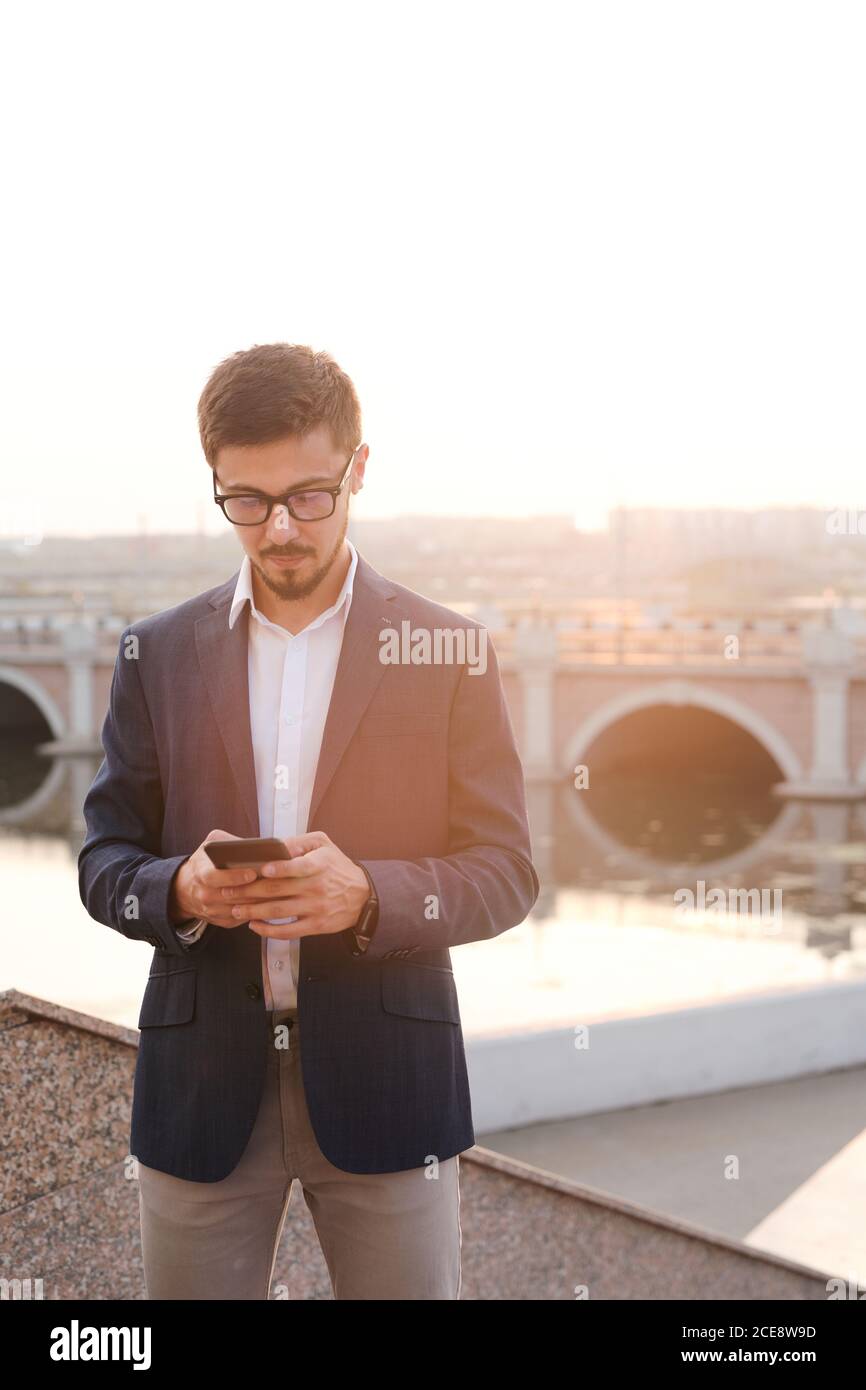 Jeune homme d'affaires sérieux barbu dans des lunettes debout contre le pont et utilisation du smartphone lors de l'envoi d'un message texte à un collègue Banque D'Images