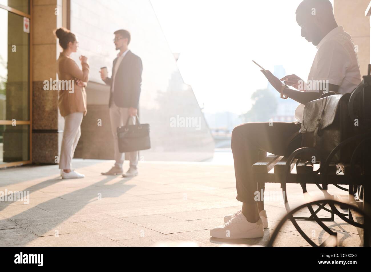 Homme noir occupé utilisant une tablette numérique et assis sur un banc pendant que les collègues boivent du café en arrière-plan Banque D'Images