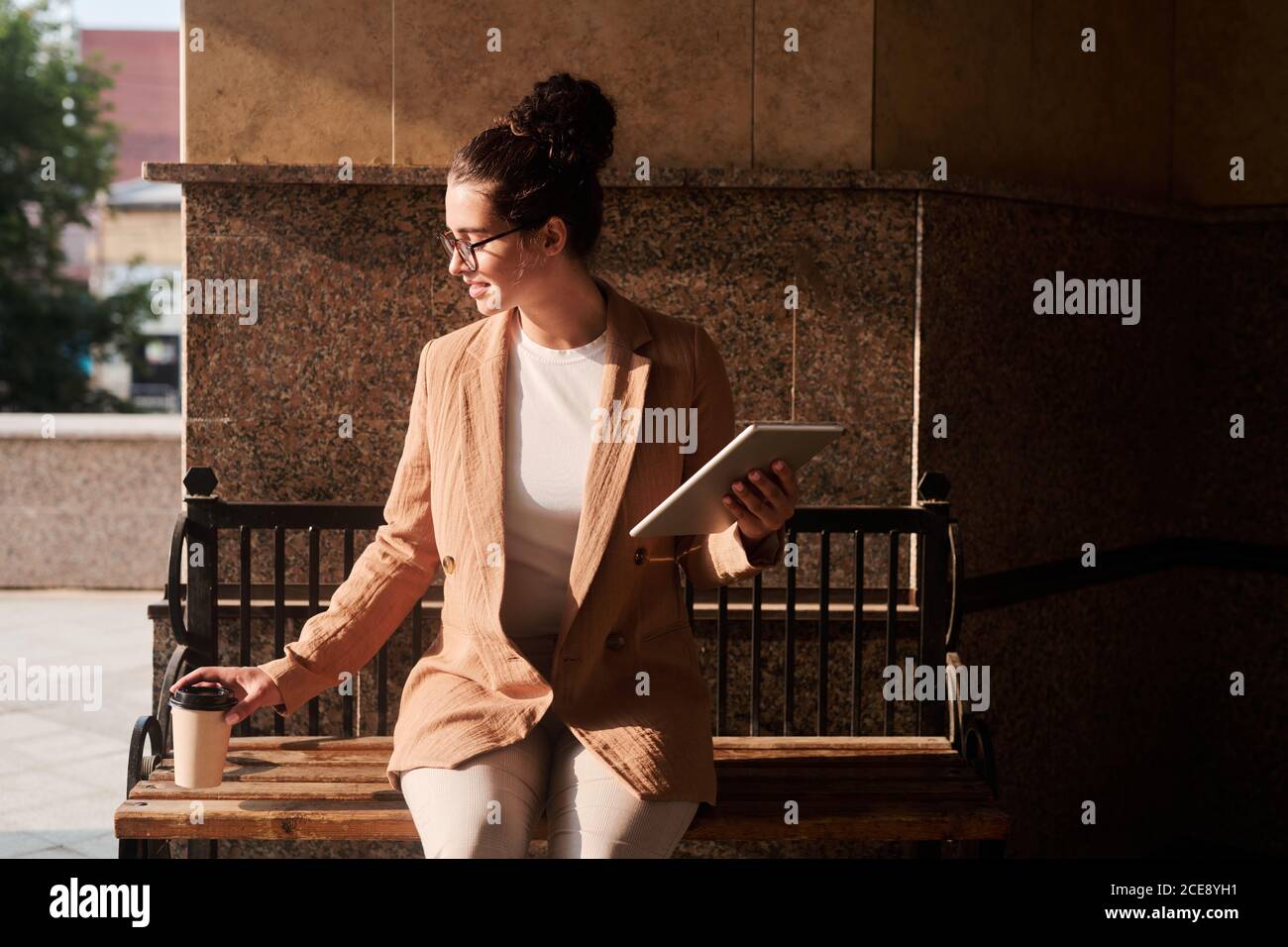 Jeune femme d'affaires souriante assise sur un banc et buvant du café lorsque vous travaillez avec une tablette à l'extérieur Banque D'Images