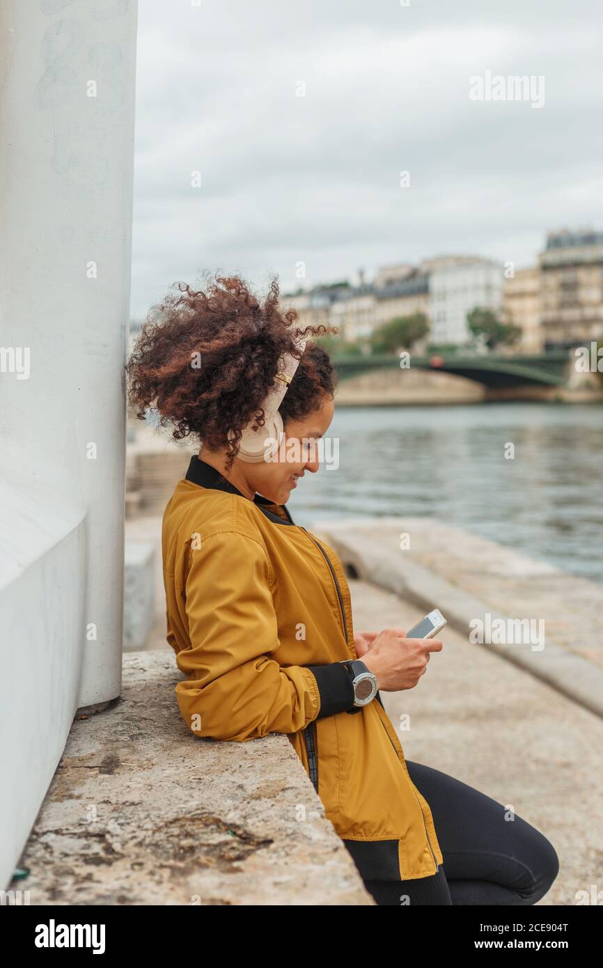 Vue latérale de l'athlète féminine ethnique en tenue active et un casque bavarde sur un téléphone portable après un entraînement en étant debout sur un remblai avec haltères près de la rivière de la ville Banque D'Images