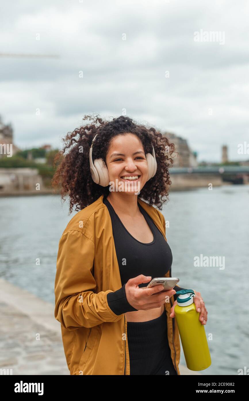 Femme athlète en tenue active et casque tenant un téléphone portable après entraînement en se tenant sur une bankment près de la rivière de la ville Banque D'Images