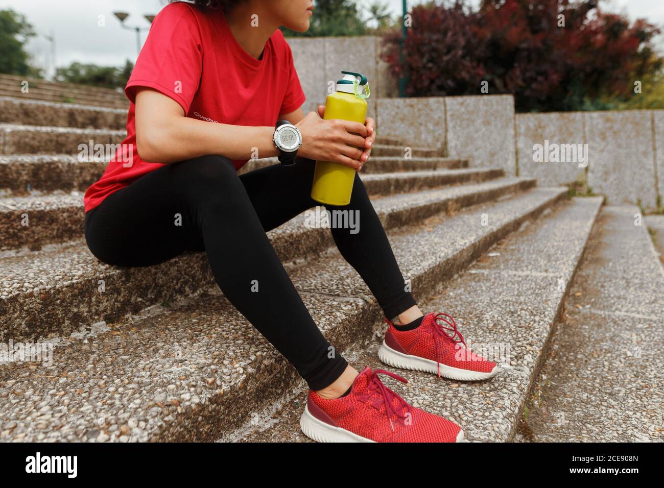Vue latérale d'une athlète féminine anonyme de sport des vêtements et une montre intelligente buvant de l'eau par thermos pendant que vous vous reposez avec les yeux fermés sur l'escalier après l'entraînement à la lumière du soleil Banque D'Images