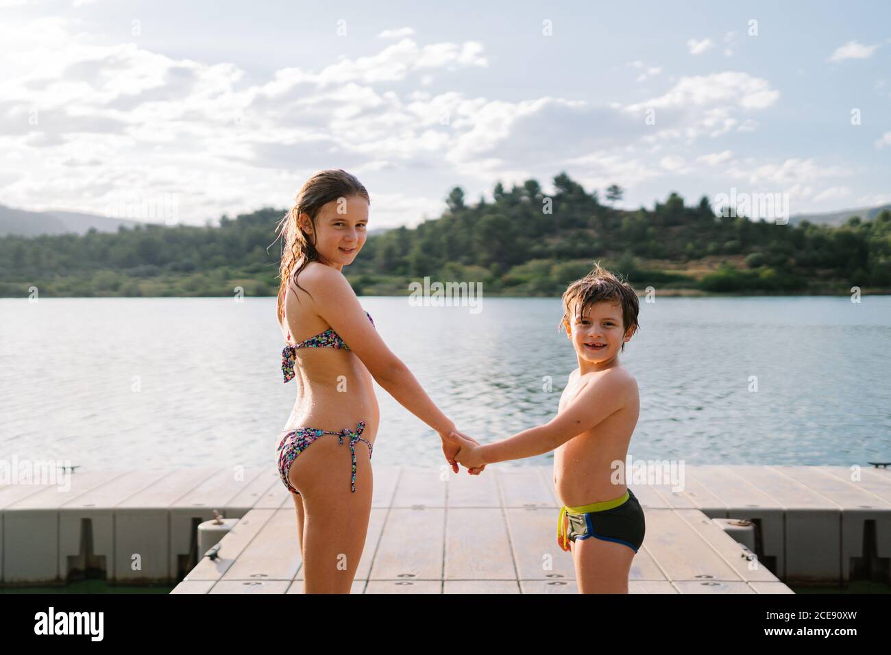 Vue latérale du frère souriant et de la sœur en maillot de bain debout  ensemble en tenant les mains sur le quai près du lac et en regardant  appareil photo tout en