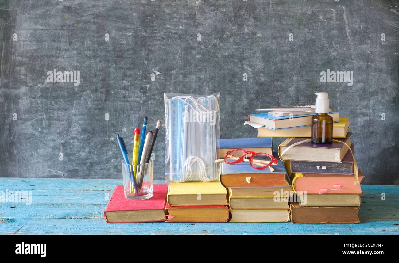 Fournitures scolaires et articles de prévention COVID 19 sur le bureau de la salle de classe avec des livres, des lunettes, des stylos sur fond de tableau noir. Retour à l'école pendant corona-viru Banque D'Images