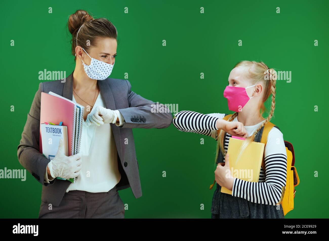 enseignante et élève moderne avec masques, sac à dos jaune et manuel accueilli avec une bosse de coude isolée sur fond vert de tableau noir. Banque D'Images