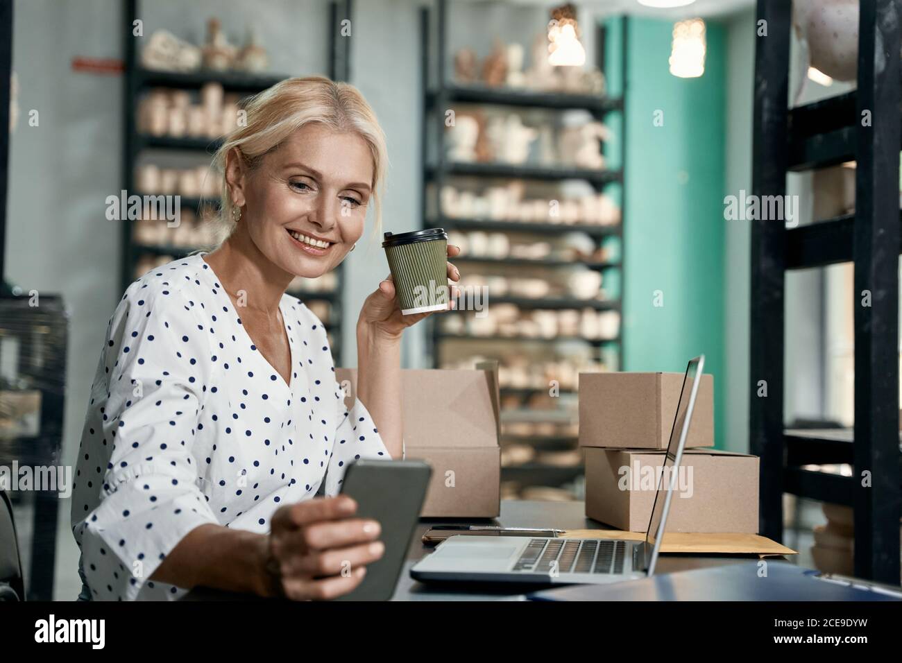 Propriétaire d'entreprise ayant réussi. Belle et heureuse femme d'affaires qui boit du café, utilise un smartphone et sourit tout en travaillant dans son studio d'art ou dans une boutique de poterie artisanale. Petite entreprise, vente au détail Banque D'Images