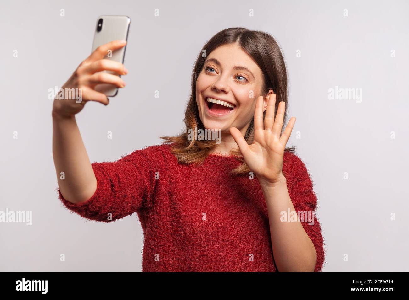 Portrait d'une fille aimable dans un chandail déchiqueté faisant des appels vidéo via un téléphone mobile et levant la main faisant des gestes bonjour, communiquant à l'aide d'Internet-activé e Banque D'Images