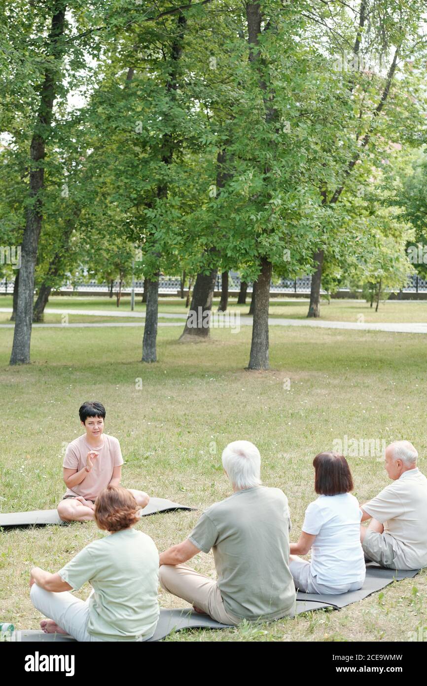Instructeur de yoga d'âge moyen assis sur un tapis dans le parc et expliquant technique de respiration pour les étudiants âgés Banque D'Images
