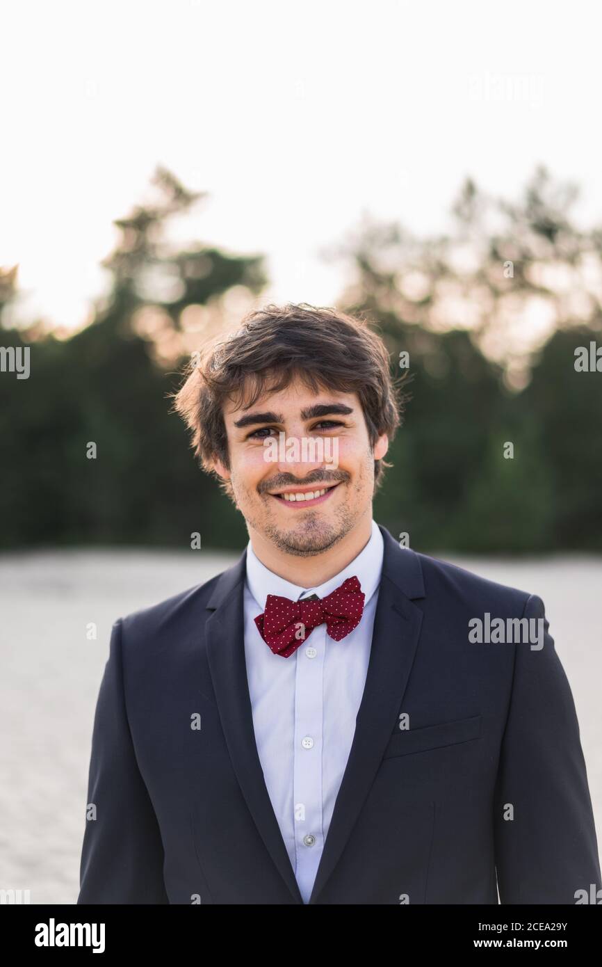 Homme adulte portant un élégant costume noir avec noeud papillon et se tenir sur la plage avec les mains dans les poches en regardant l'appareil photo Banque D'Images