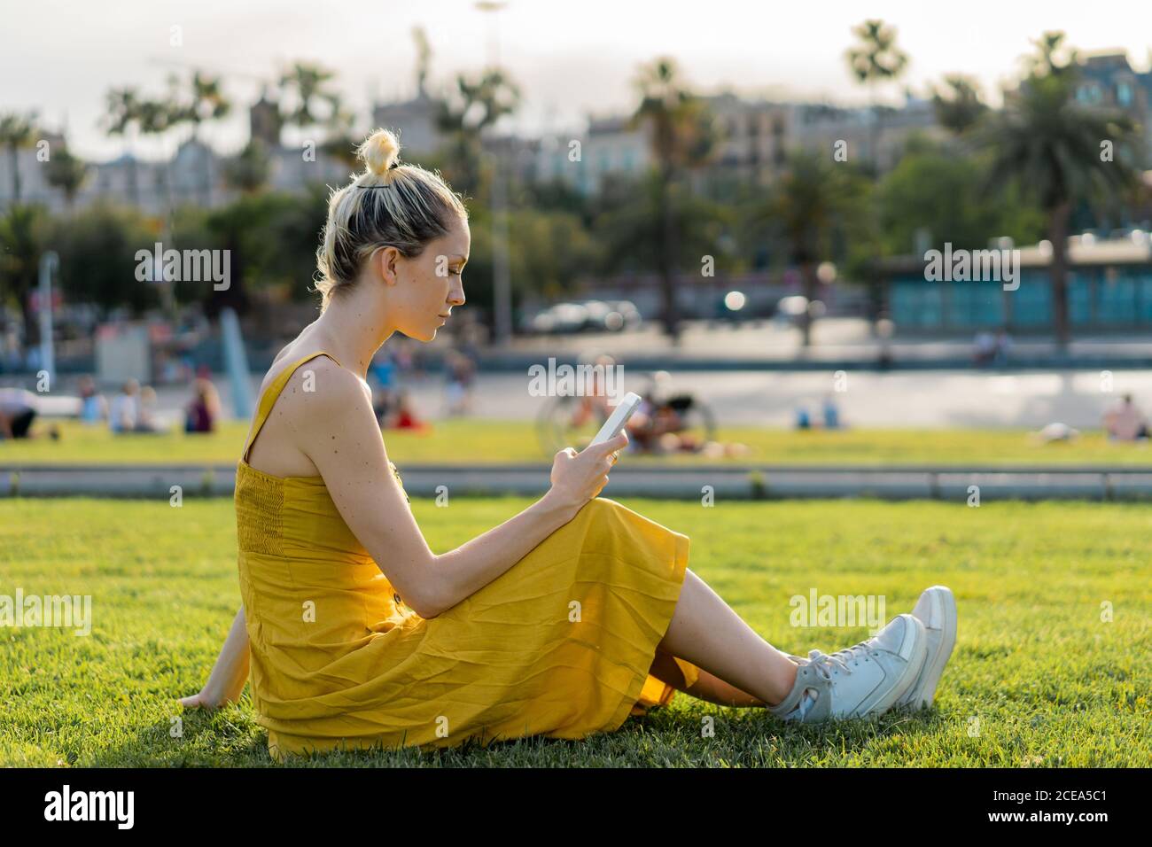 Vue latérale d'une jolie femme blonde avec cheveux attachés robe d'été jaune longue et baskets décontractées sur pelouse avec le téléphone mobile appuyé sur le bras Banque D'Images