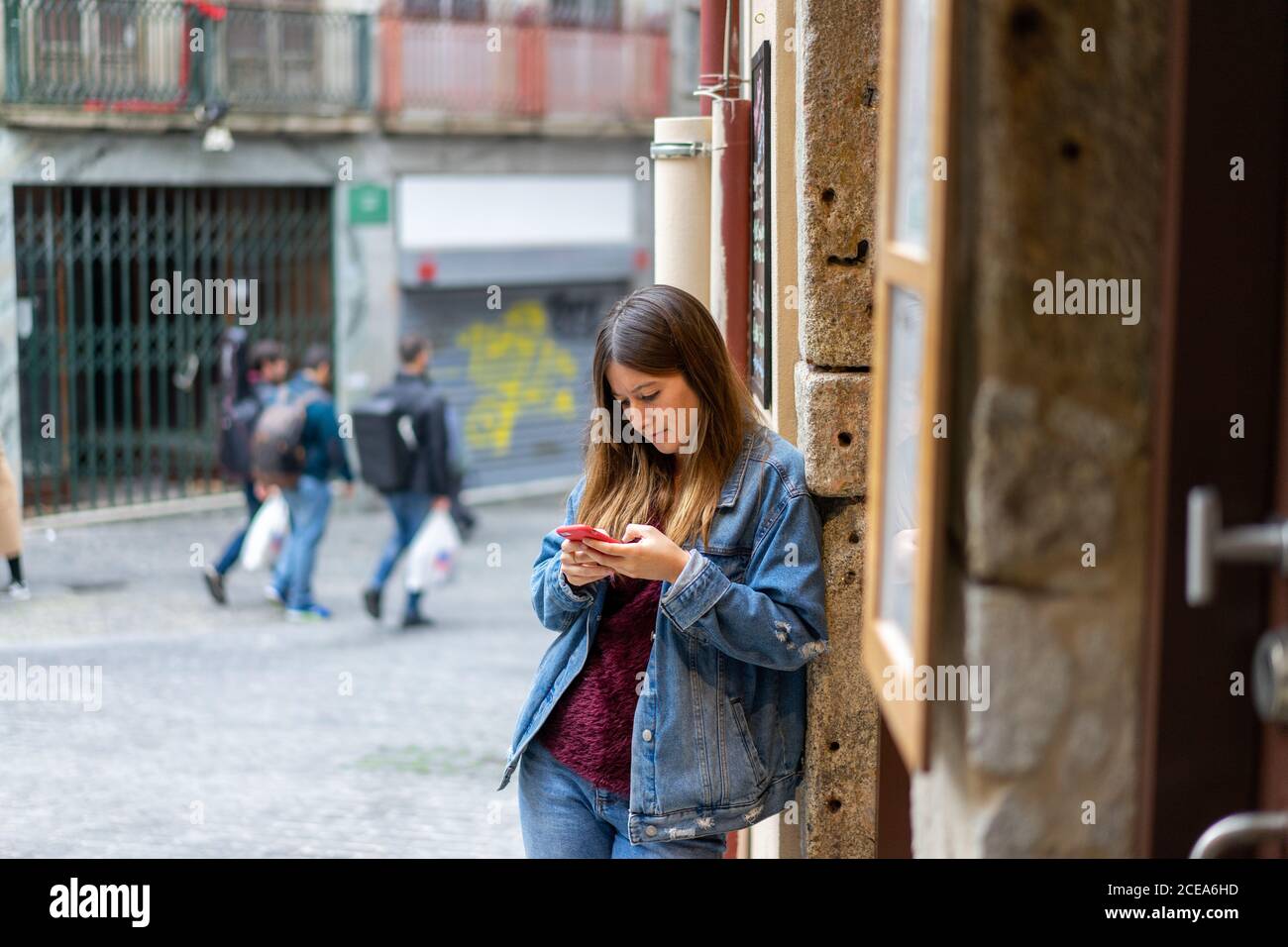 Belle femme en jean blouson tenant un téléphone mobile rouge et debout près du mur dans la rue à Porto, Portugal Banque D'Images