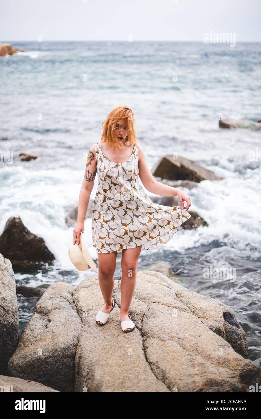 Vue arrière de la femme tatouée dans un chapeau debout sur des pierres avec les mains à part et regardant l'océan Banque D'Images