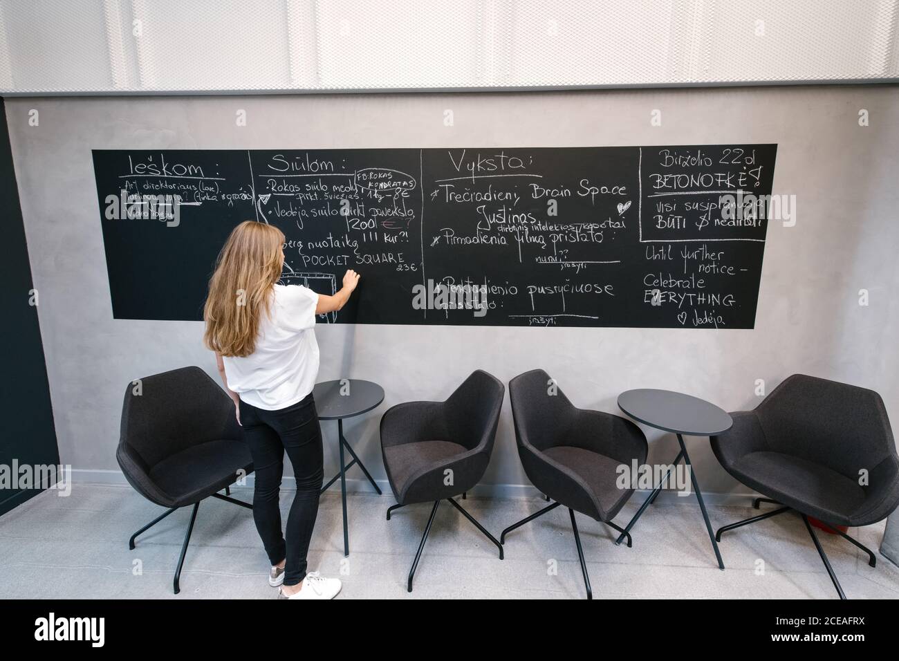 Vue arrière d'une femme décontractée qui écrit sur un tableau noir dans le hall d'un bureau moderne avec des chaises et des tables à proximité Banque D'Images