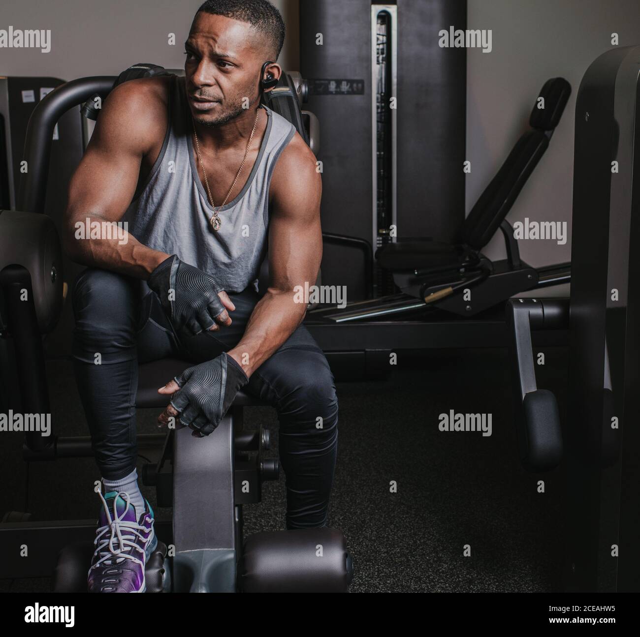 Séduisant Afro-américain gars dans sportswear à l'écoute de la musique dans ecouteurs et regarder loin en étant assis sur la machine d'exercice salle de sport moderne Banque D'Images