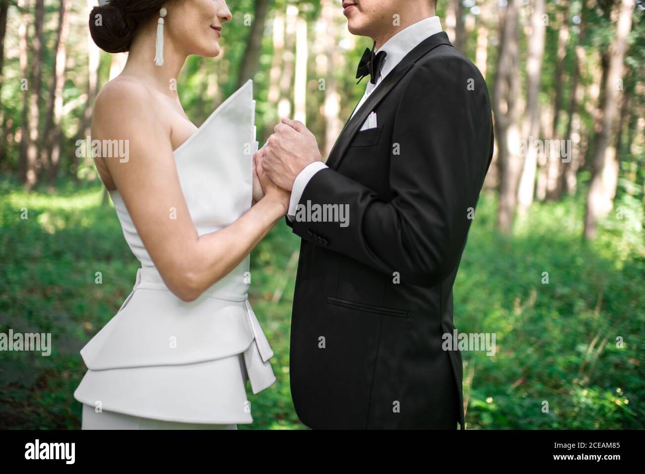 Vue latérale d'un jeune marié méconnu et d'une jeune mariée qui se touche les mains de l'autre tout en se tenant sur un arrière-plan flou de belle forêt le jour ensoleillé Banque D'Images