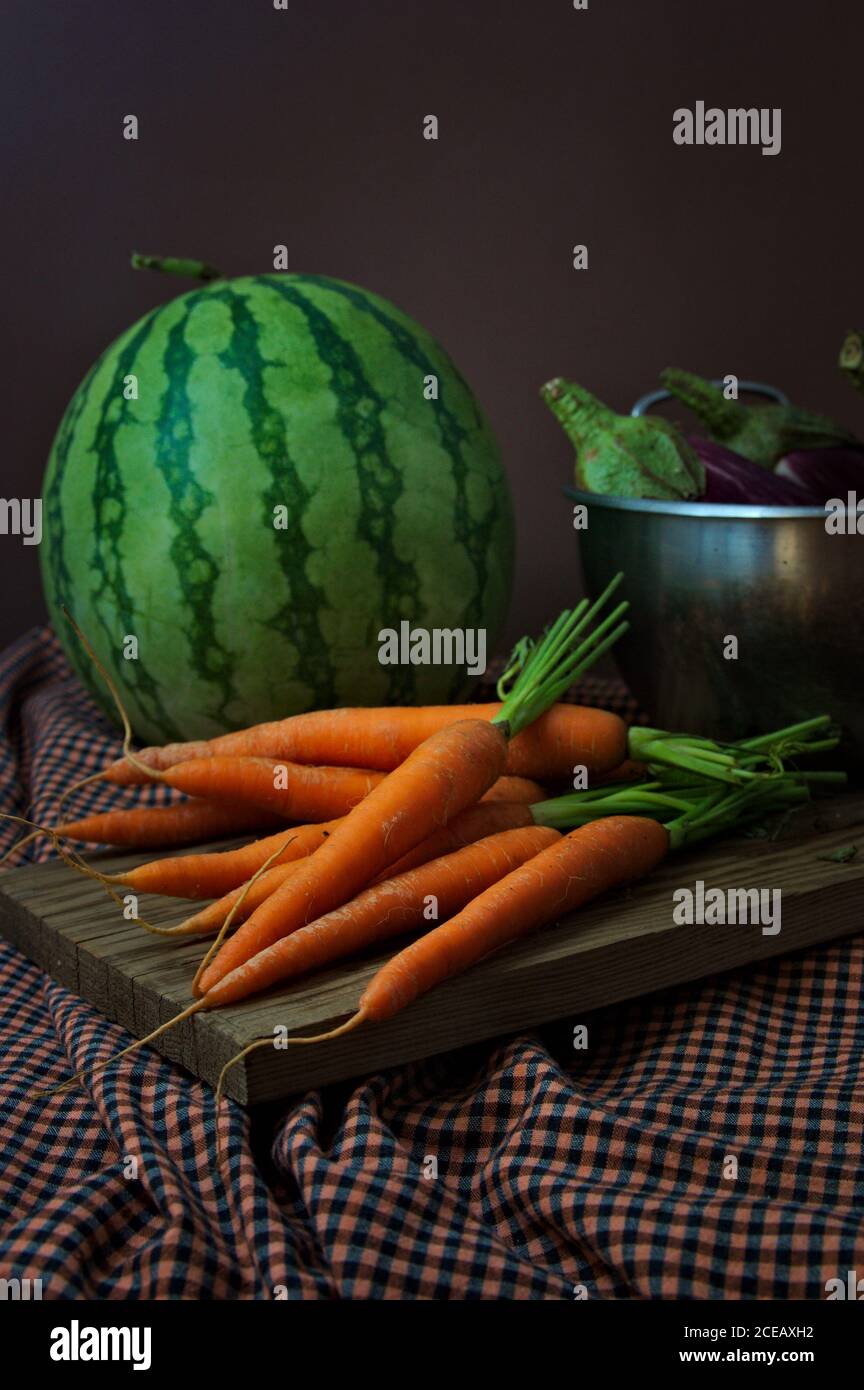 La vie verticale des fruits où les aubergines, les carottes apparaissent sur une planche de bois et une pastèque rayée avec un fond sombre Banque D'Images