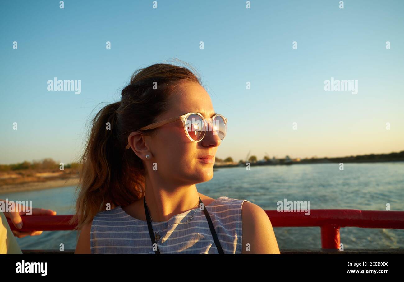Charmante femme aux cheveux bruns et aux lunettes de soleil vêtue d'une chemise rayée grise sur bateau avec côte floue et ciel bleu sur fond Banque D'Images