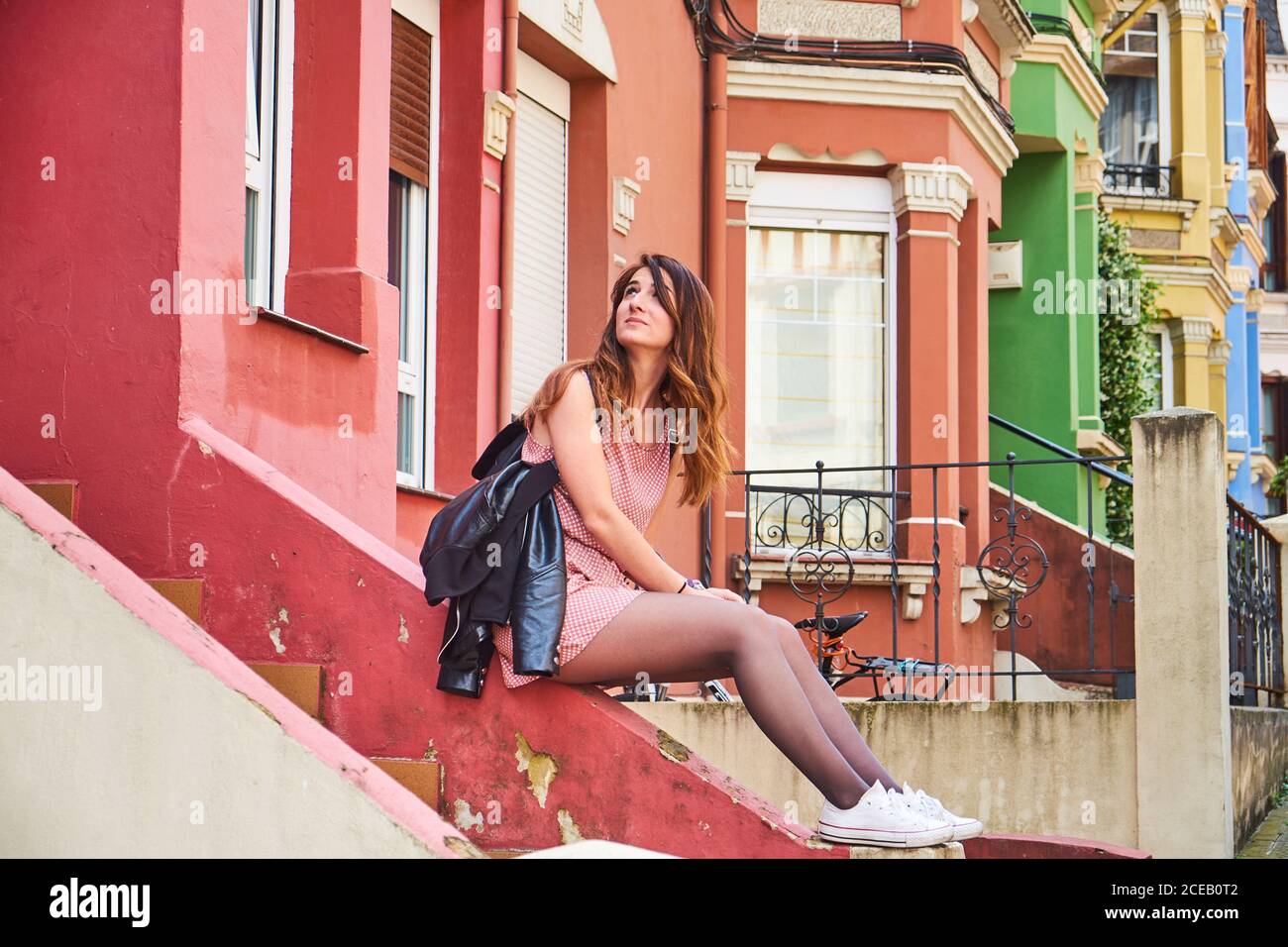 Vue latérale de la jeune belle femme en robe courte assise sur l'escalier du bâtiment lumineux et regardant la façade Banque D'Images