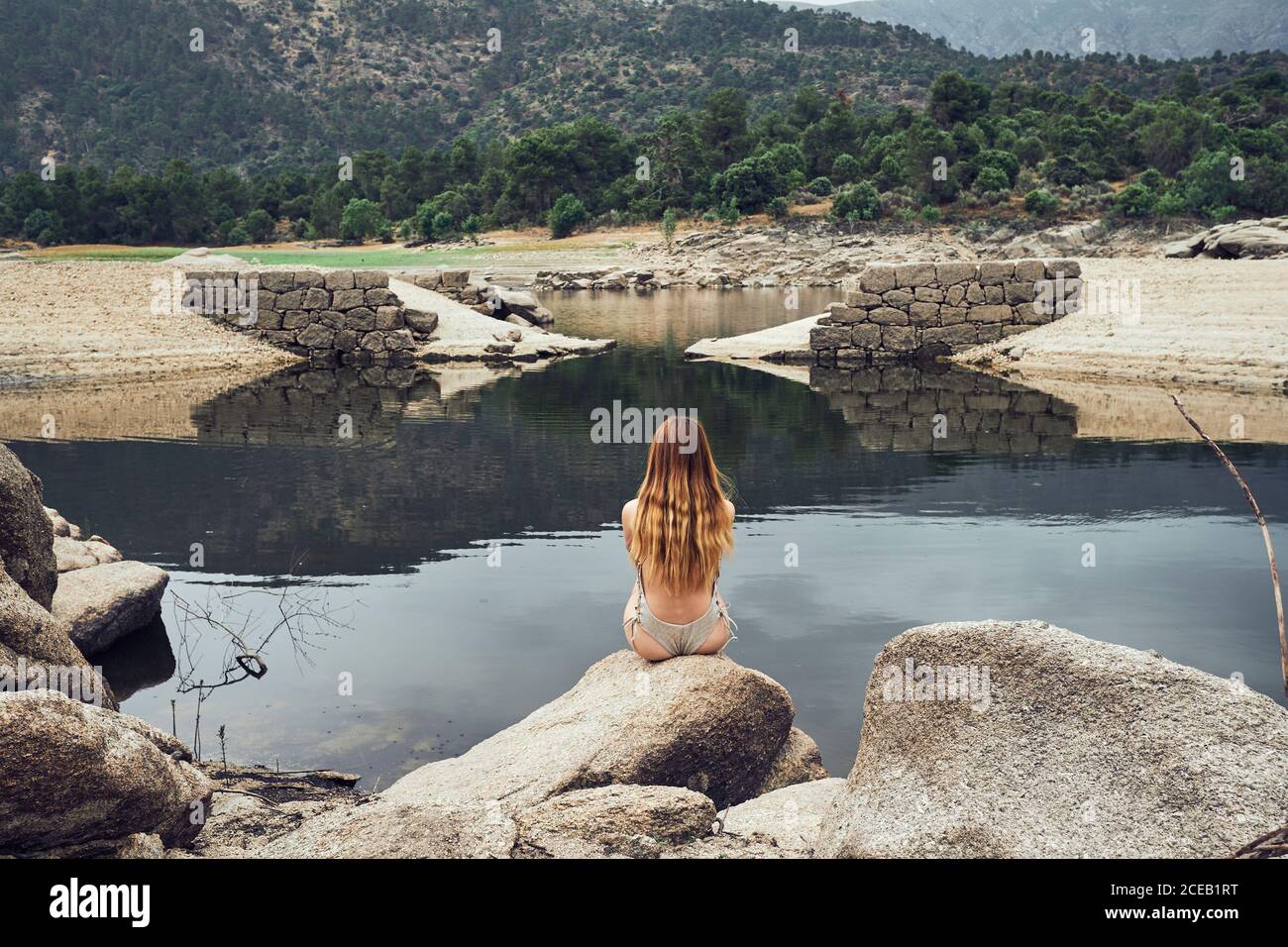 Vue arrière de la jeune femme avec de longs cheveux en maillot de bain assis sur une grande pierre et regardant le lac STILL sur le fond des arbres verts et des montagnes Banque D'Images
