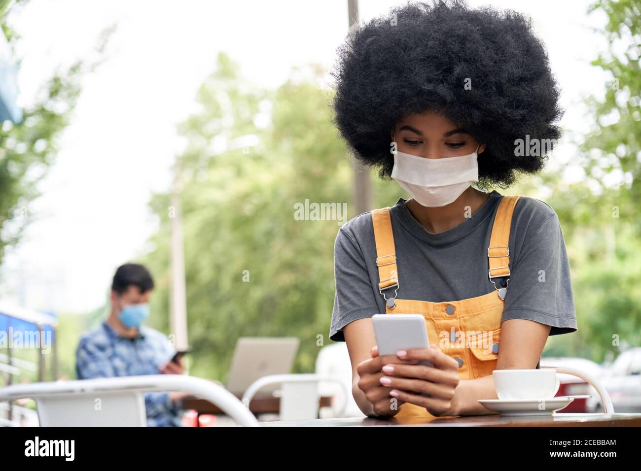 Une femme africaine portant un masque facial avec un téléphone s'assoit à la table dans un café extérieur. Banque D'Images