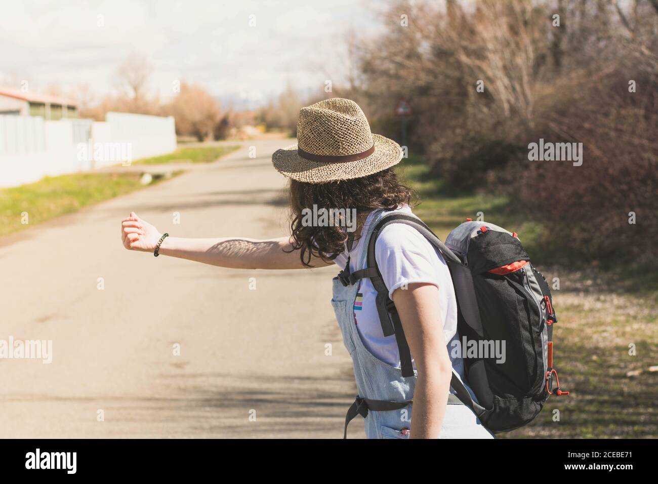 Vue latérale d'une brunette décontractée en chapeau et avec sac à dos randonnée sur une route rurale ensoleillée avec le pouce vers le haut Banque D'Images
