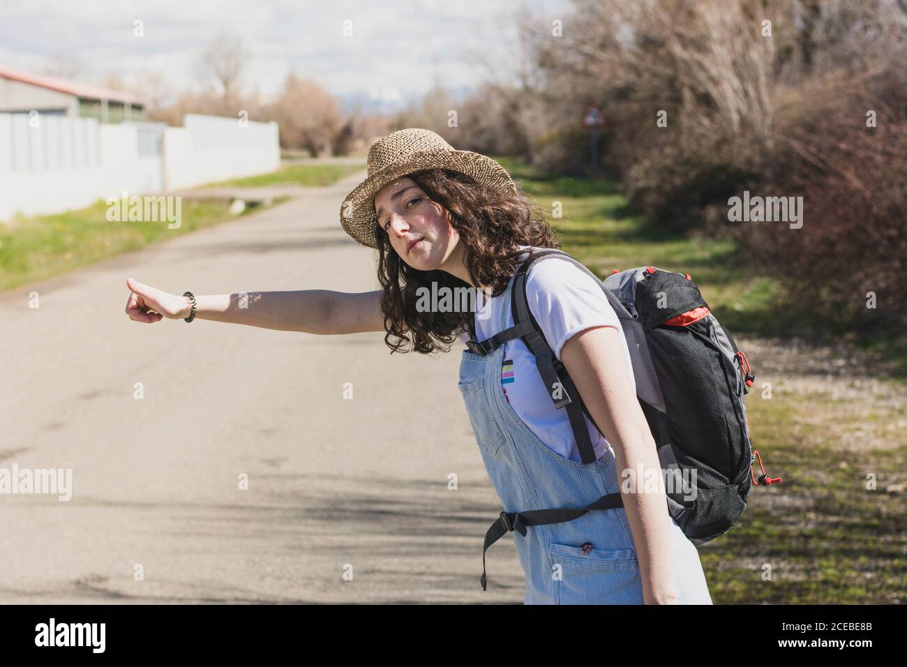 Vue latérale d'une brunette décontractée en chapeau et avec sac à dos randonnée sur une route rurale ensoleillée avec le pouce vers le haut Banque D'Images