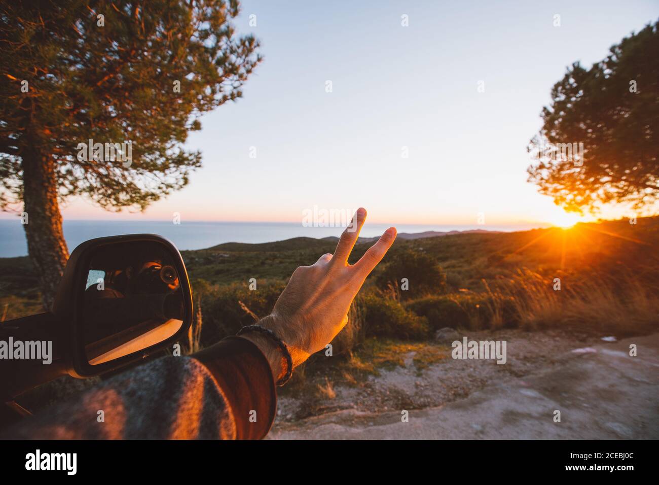 Main de l'homme anonyme montrant le V-sign près du miroir d'aile contre campagne majestueuse au beau coucher du soleil Banque D'Images