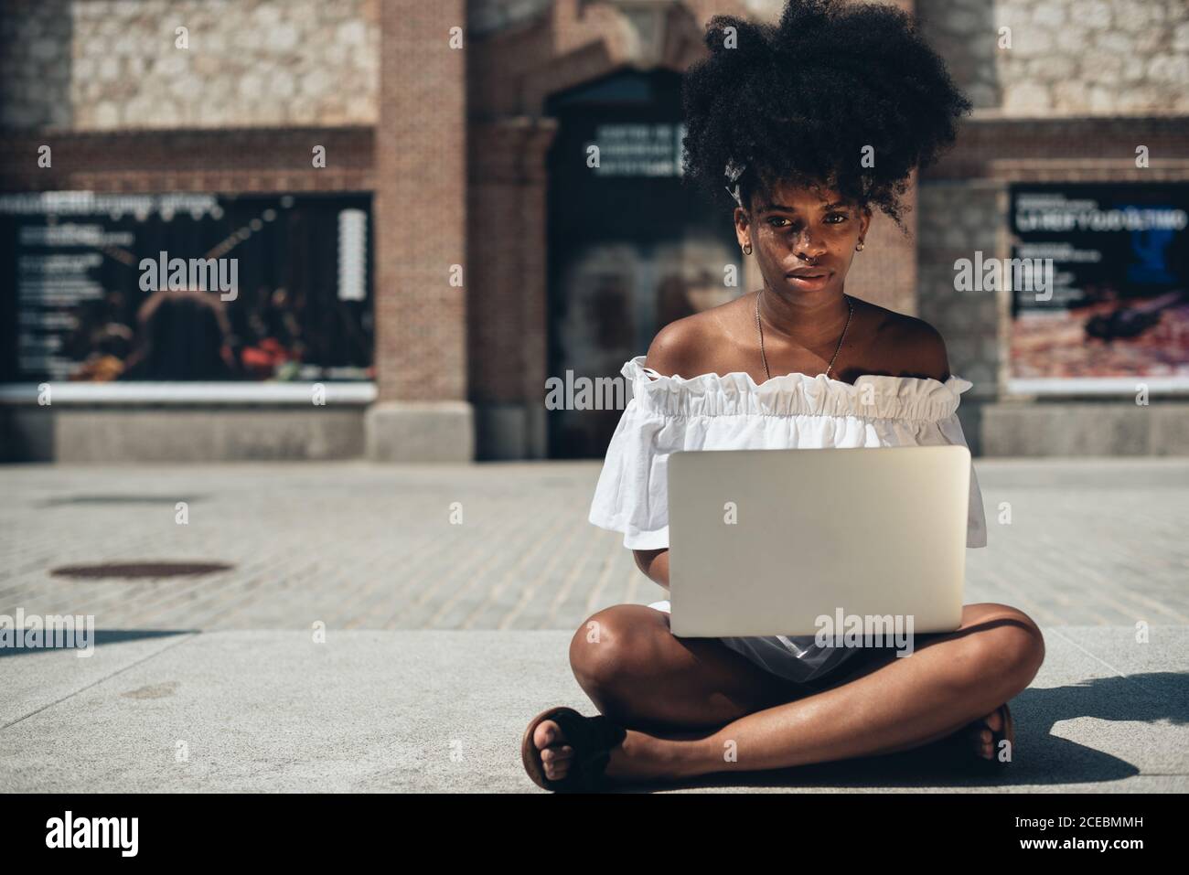 Jolie femme afro-américaine moderne en robe blanche assise sur le trottoir avec les jambes croisées et en utilisant un ordinateur portable au soleil Banque D'Images