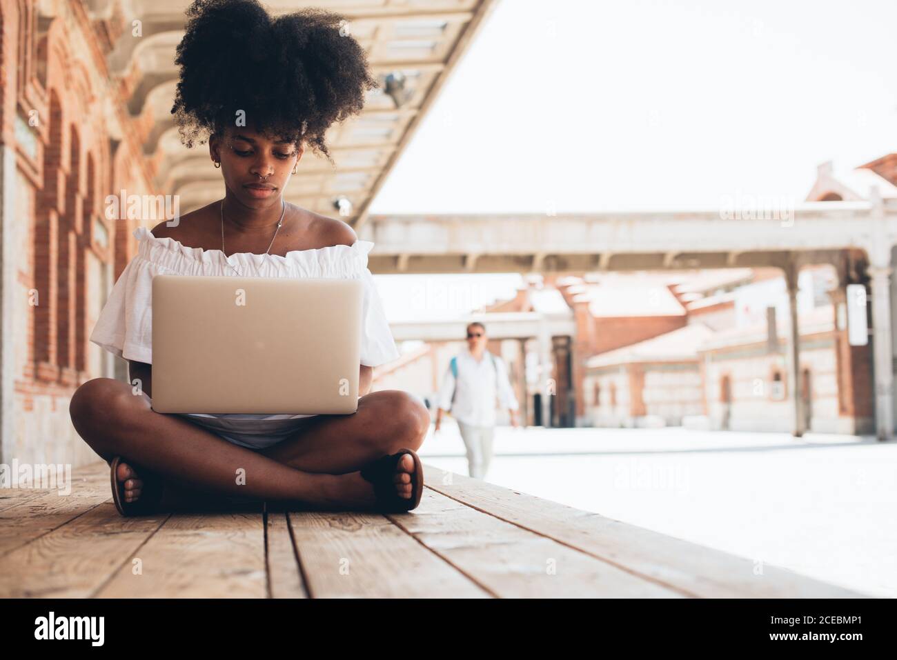 Jolie femme afro-américaine moderne en robe blanche assise sur le trottoir avec les jambes croisées et en utilisant un ordinateur portable au soleil Banque D'Images