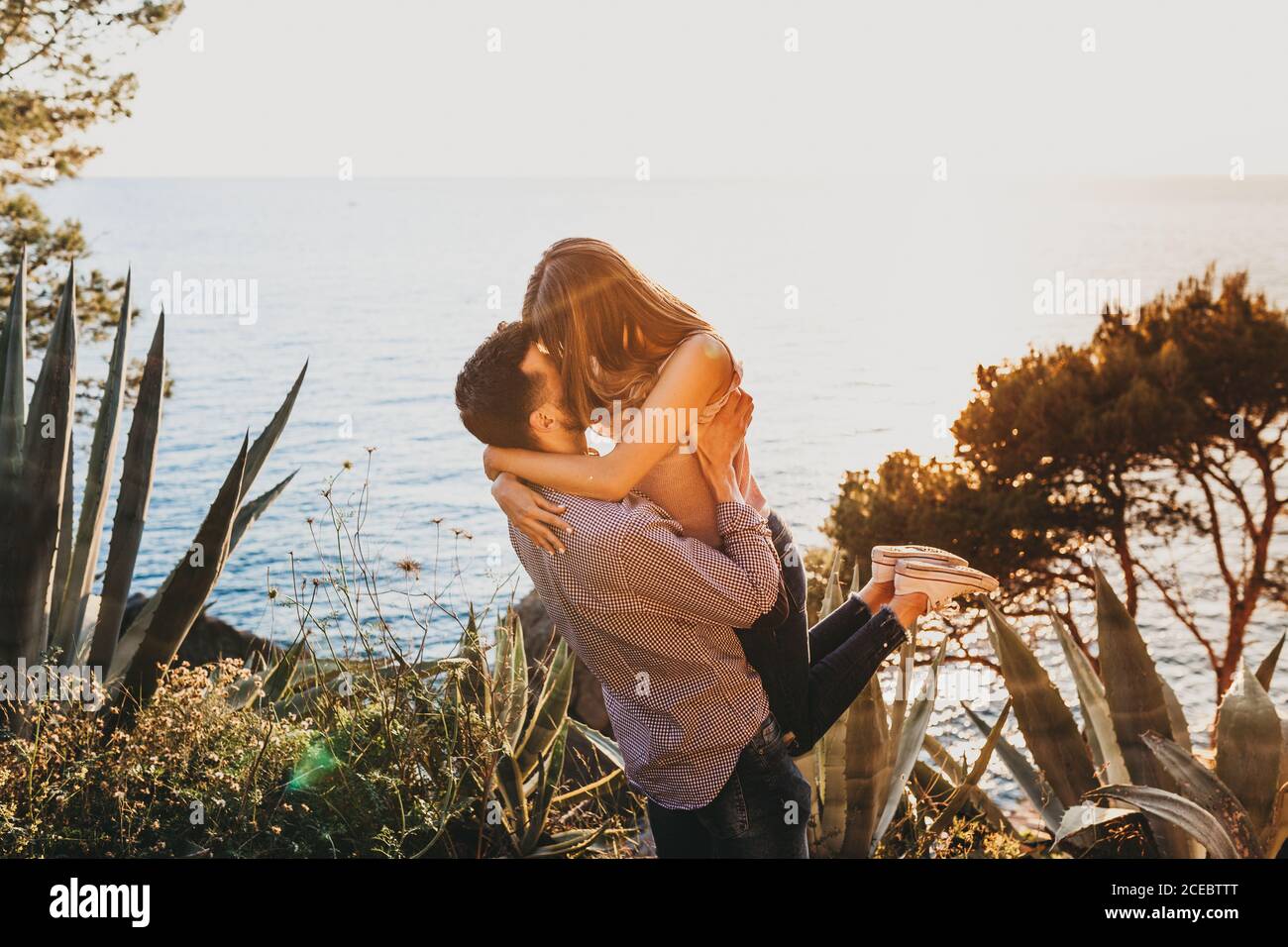 Homme en vêtements d'été tenant dans les bras jeune femme embrassant et l'embrassant debout sur la colline avec des arbres et des plantes exotiques sur la côte de l'océan le jour ensoleillé d'été Banque D'Images
