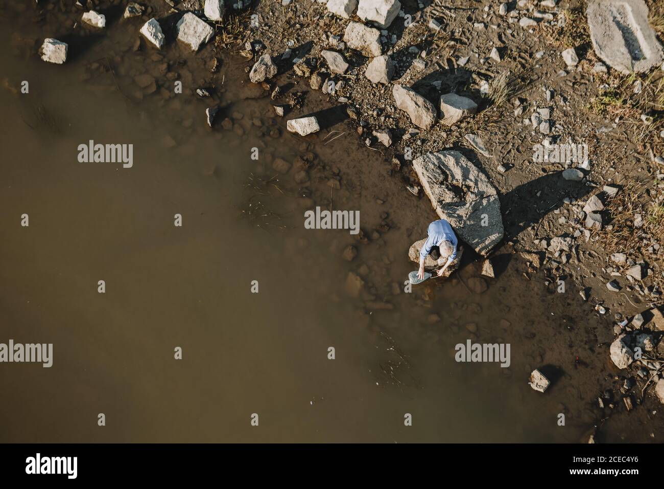 Homme assis sur la pierre dans l'eau des bancs sur la rive brune de la rivière Tisza brune en Hongrie à Szeged, prenant l'eau dans la bouteille d'eau Banque D'Images