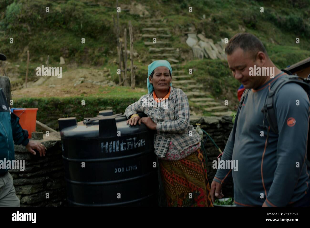 Un homme portant un sac à dos marchant sur un chemin rural, passant devant une femme debout à côté d'un grand réservoir d'eau en plastique au village écotouristique de Bhanjyang sur la région de montagne de Panchase, Gandaki Pradesh, Népal. Banque D'Images Un homme portant un sac à dos marchant sur un chemin rural, passant devant une femme debout à côté d'un grand réservoir d'eau en plastique au village écotouristique de Bhanjyang sur la région de montagne de Panchase, Gandaki Pradesh, Népal. Banque D'Images