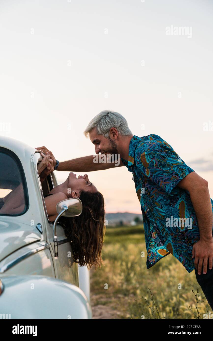 Vue latérale d'une femme assise à l'intérieur d'une voiture penchée tête dehors et regardant un homme debout au-dessus et souriant heureux en été Banque D'Images