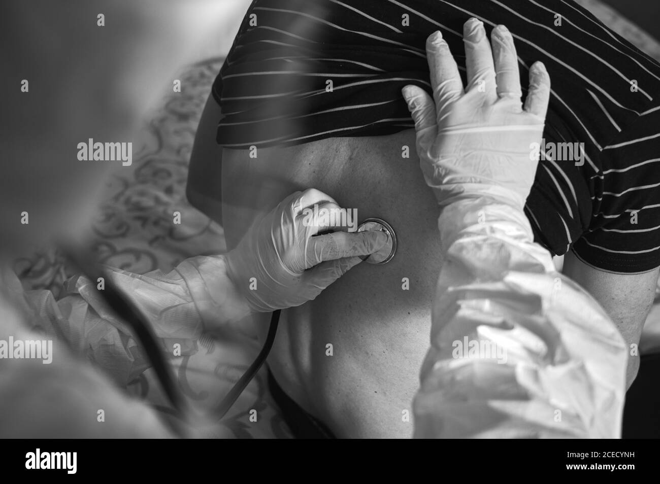 Prise de vue rapprochée de la main du médecin en combinaison de protection PPE examinant un patient mâle âgé à la maison à l'aide d'un phonendoscope, en vérifiant son dos. Image en noir et blanc. Concept COVID-19 Banque D'Images