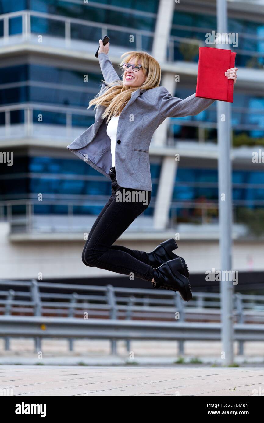 Belle blonde avec des lunettes en veste grise et jeans noirs sourire et sauter avec un sac rouge et un téléphone dans les mains Banque D'Images
