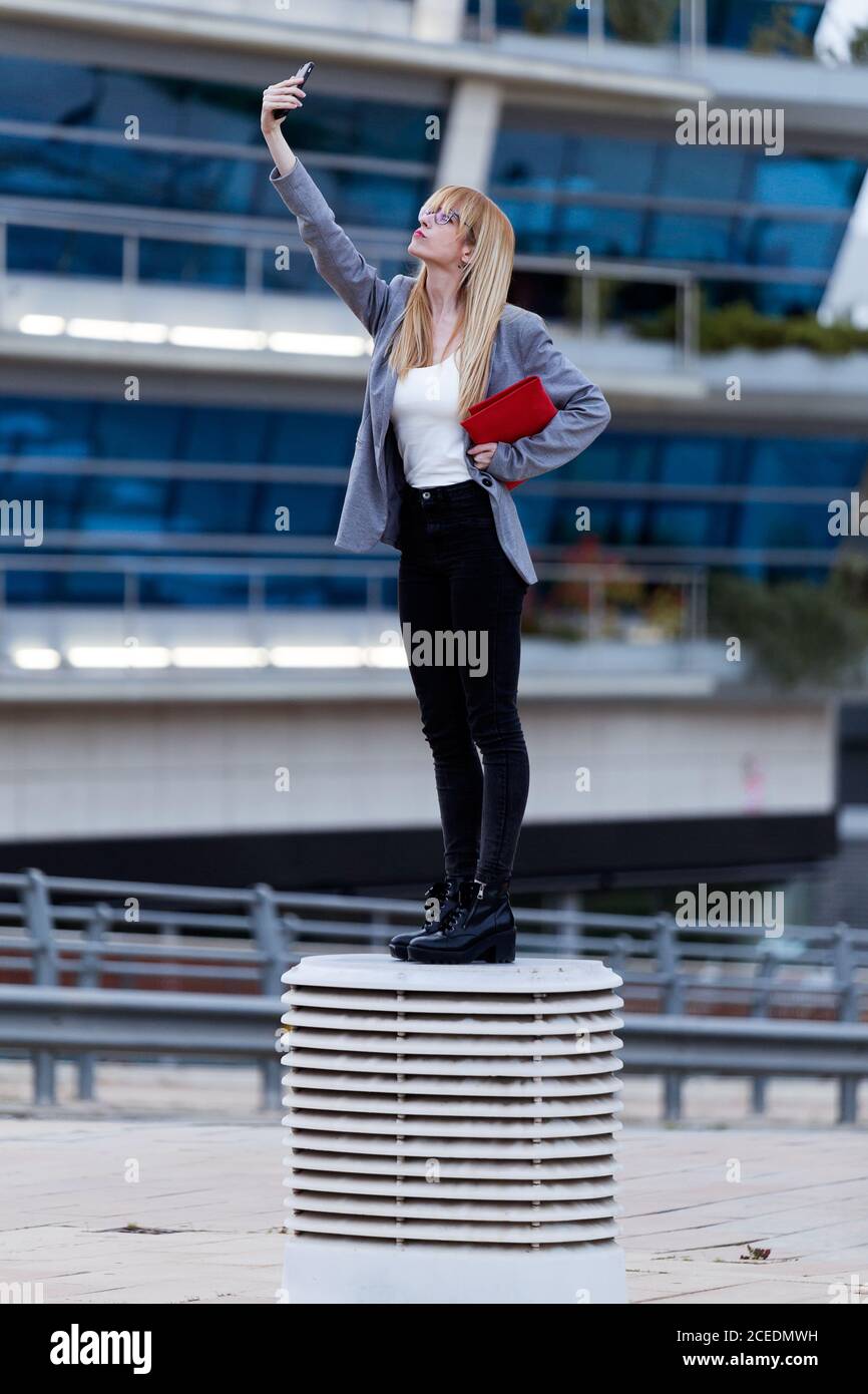 Belle femme en blouson gris et jeans noirs faisant photo sur téléphone avec rouge à lèvres Banque D'Images