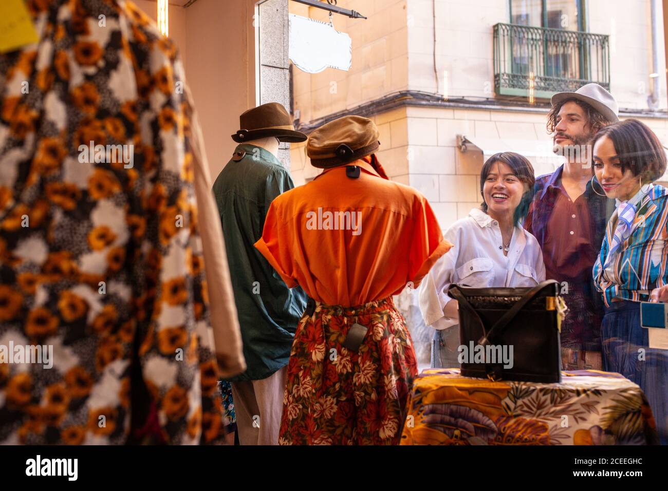 Jeunes hommes et femmes souriant et regardant les vêtements à l'intérieur un petit magasin se tenant dans la rue près de la fenêtre de présentation Banque D'Images