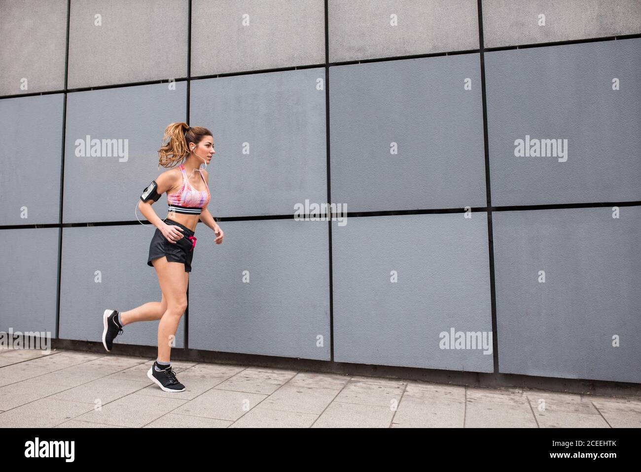 Vue latérale d'une femme musclée en short et haut écoutant de la musique tout en courant dans une rue vide contre un bâtiment moderne Banque D'Images