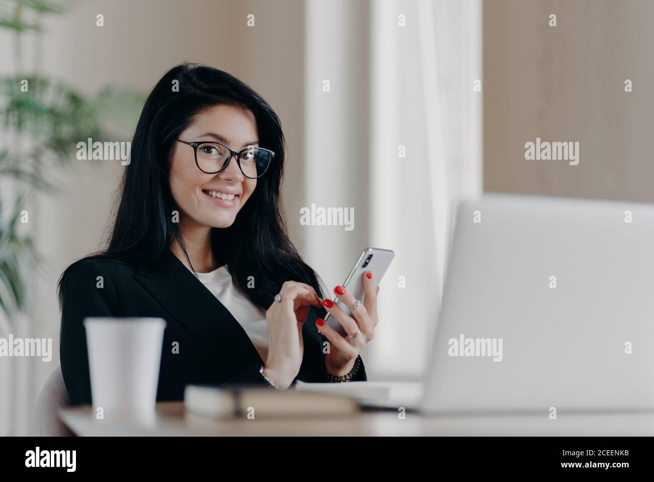 Prise de vue horizontale des messages européens de la femme brunette heureuse sur smartphone moderne, donne une consultation en ligne, sourit positivement, porte des lunettes et pour Banque D'Images