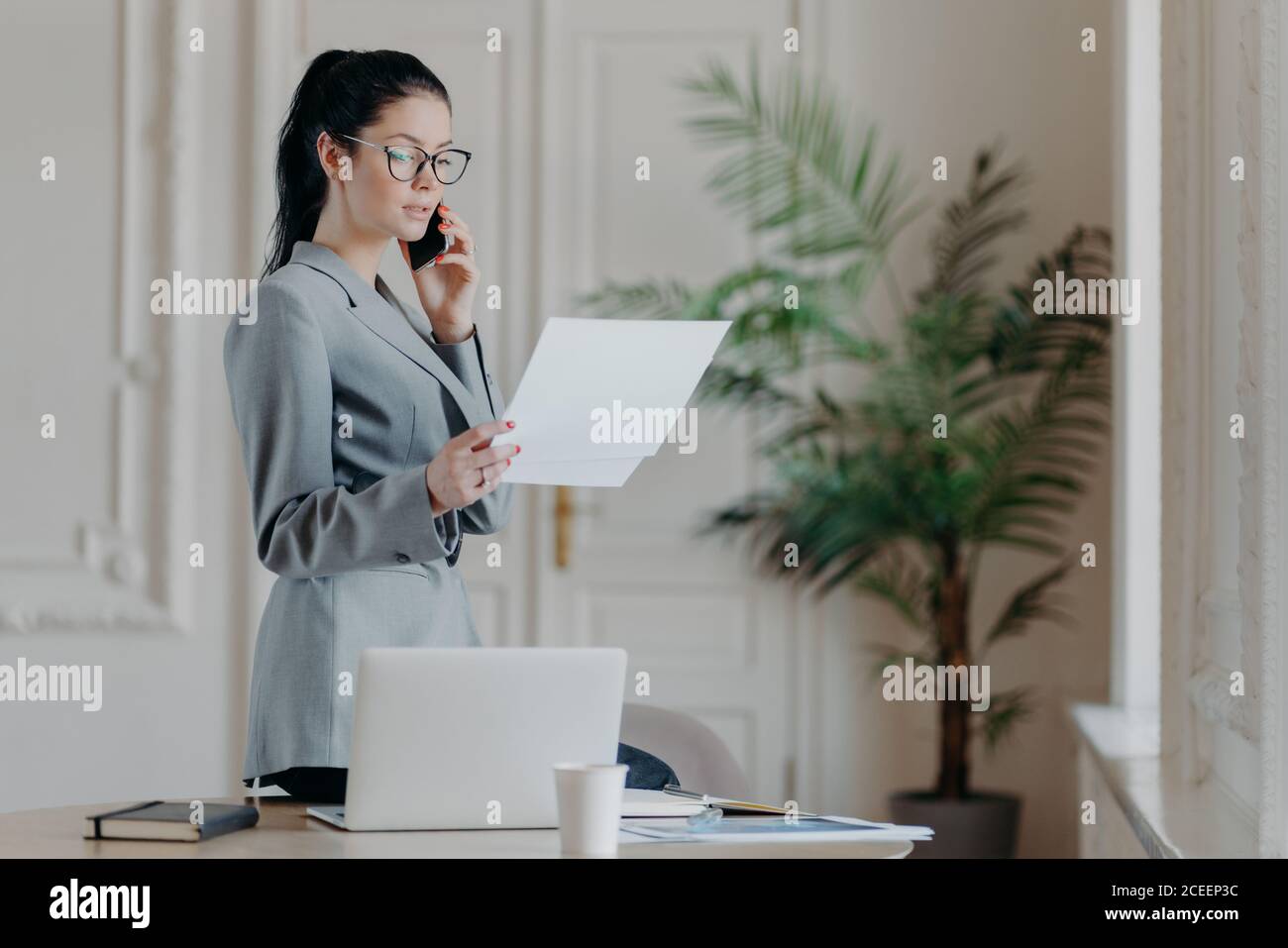 Une employée brune sérieuse se tient près d'un bureau, tient des documents papier, parle via un smartphone, porte des vêtements et des lunettes formels, discute de busi Banque D'Images