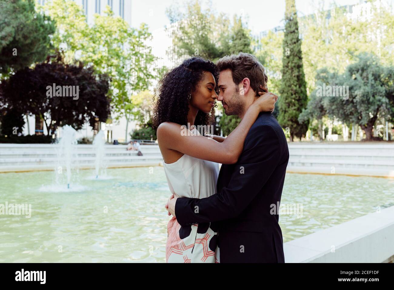 Vue latérale de charmante femme noire et beau homme caucasien s'embrassant tout en se tenant près de la belle fontaine dans le parc Banque D'Images