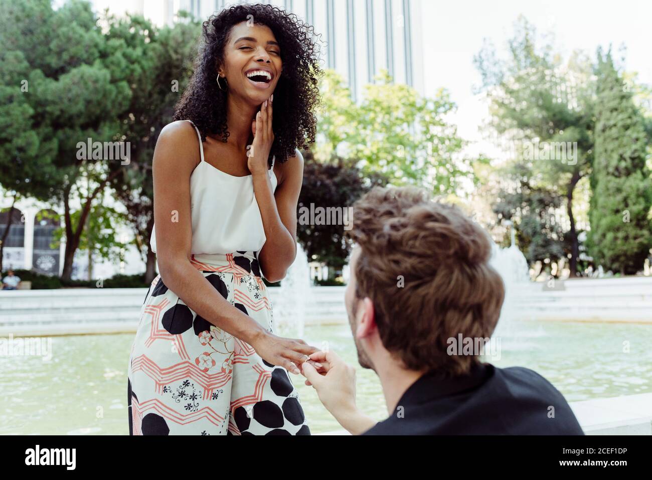Homme caucasien mettant l'anneau d'engagement sur le doigt de la femme noire tout en lui proposant près de la fontaine dans le parc Banque D'Images