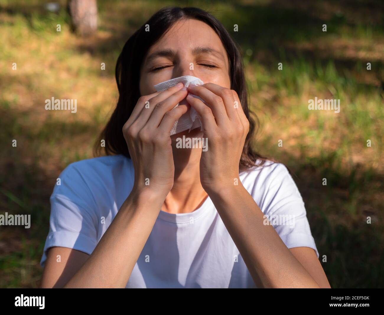Femme aux yeux fermés souffrant d'herbe de pollen allegry éternue dans un papier mouchoir un jour ensoleillé dans le parc. Concept de nez de course allergique. Banque D'Images