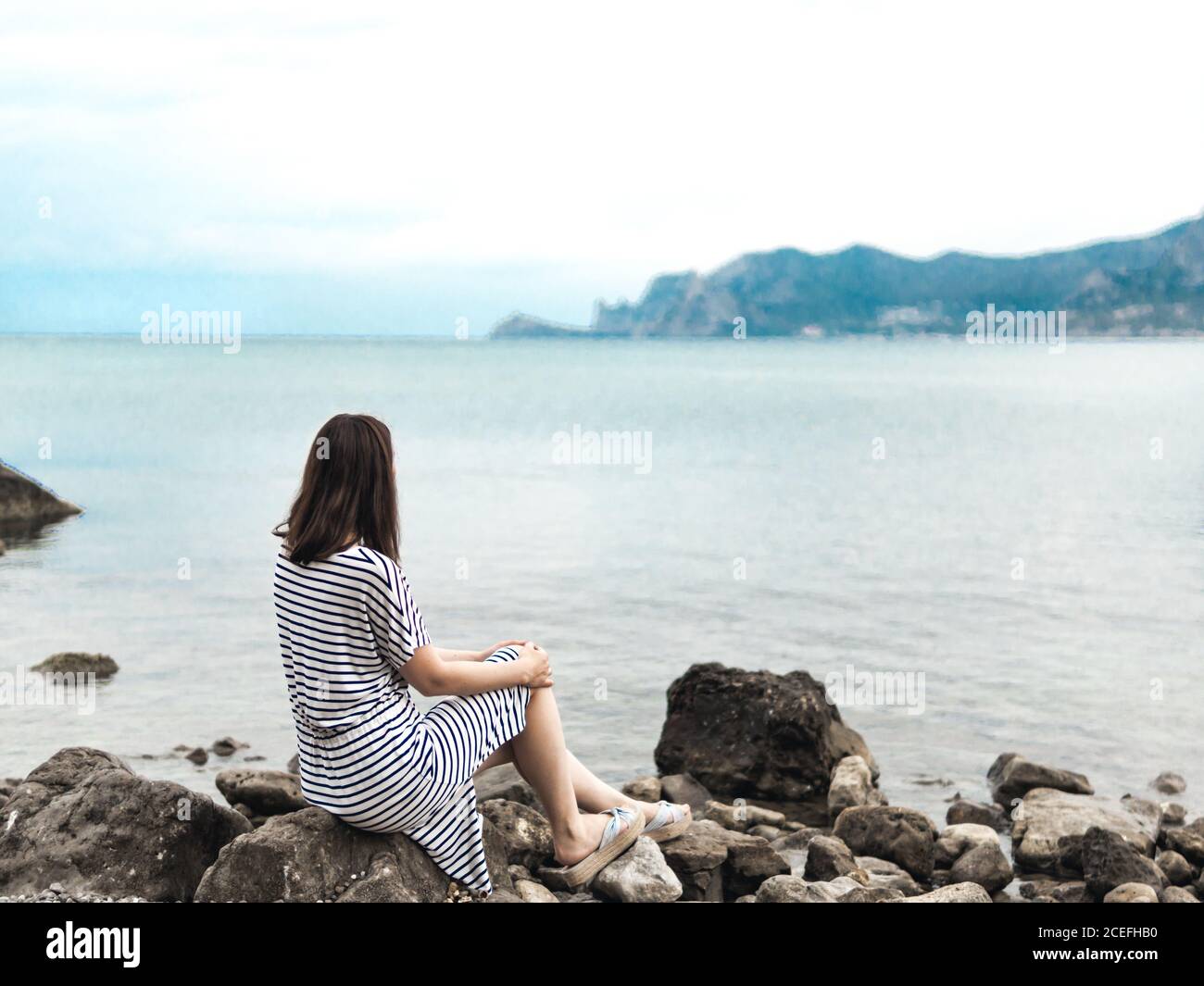 Vue arrière d'une femme en robe rayée, assise seule sur des rochers de mer, en regardant loin dans les rêves Banque D'Images