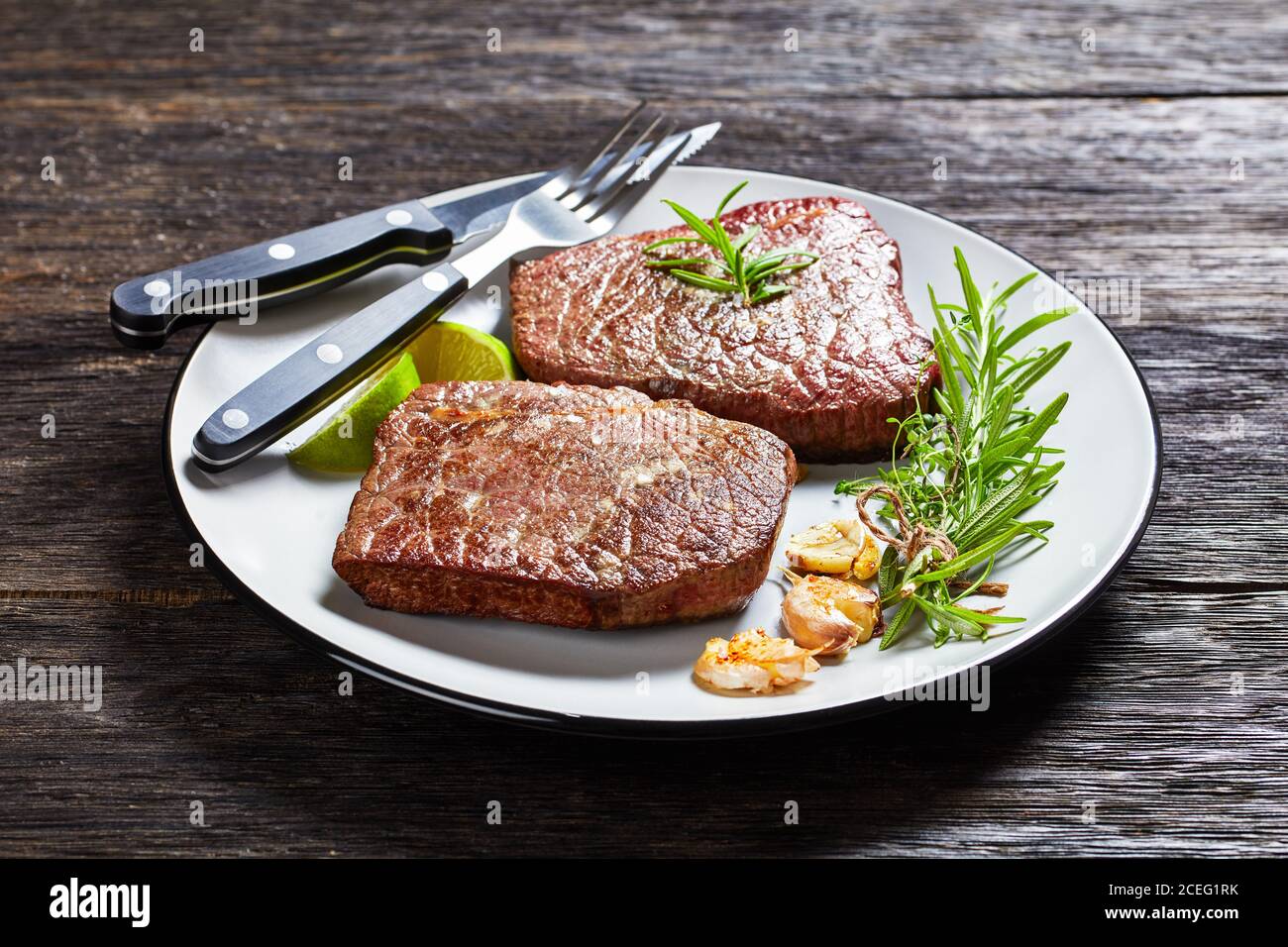 gros plan de steaks de bœuf frits au romarin, à l'ail et à la chaux sur une assiette avec fourchette et couteau sur une table en bois sombre, vue horizontale depuis le dessus Banque D'Images